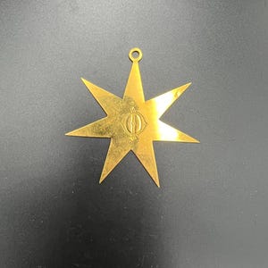 以下が含まれることがあります： 金色の八芒星型のペンダント。上部に小さなループが付いており、吊り下げることができます。星の中心には詳細なエンブレムがあります。ペンダントはおそらく金属製で、光沢のある仕上げです。背景は暗い色です。
