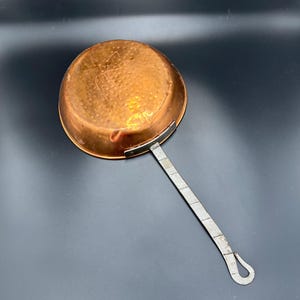 Puede incluir: Una pequeña cacerola de cobre con un mango largo de metal remachado. El interior de la cacerola tiene una textura martillada y el cobre tiene un tono marrón rojizo cálido. El mango tiene un lazo en el extremo para colgar. Utensilios de cocina.