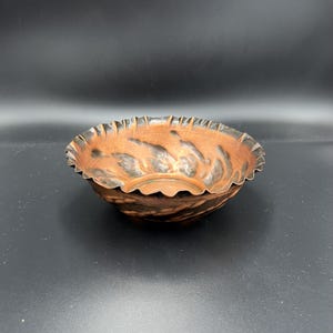 Könnte beinhalten: Eine kleine, dekorative Schale mit gerüschtem Rand. Die Schale hat eine kupferfarbene Außenseite mit dunklen, wirbelnden Mustern. Der Rand hat ein gewelltes Design. Die Schale steht vor einem dunklen Hintergrund.