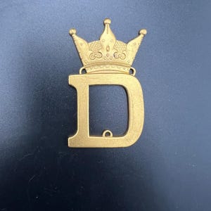 以下が含まれることがあります： 王冠が上に乗った文字「D」のゴールドカラーのメタルチャーム。王冠には複雑なディテールと3つの小さなポイントがあります。チャームの「D」の下部には小さなループが付いています。