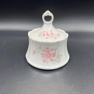 Op de afbeelding: Witte keramische suikerpot met deksel, versierd met een roze en grijs bloemmotief. De kom heeft een uitlopende vorm en een handvat op het deksel. Een klassiek, elegant ontwerp.