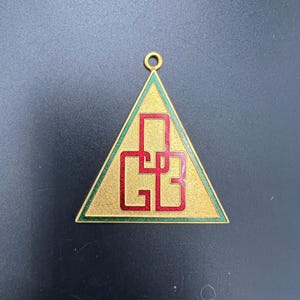 May include: Gold-toned triangular pendant with a green border and red lettering. The pendant features the letters "GB" in a stylized font. A small loop is at the top for attaching to a chain or cord.