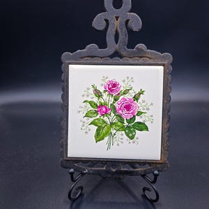 Pode incluir: Um descanso de panela decorativo com um design floral. O descanso de panela apresenta um azulejo branco com um buquê de rosas cor-de-rosa, folhas verdes e gipsofila branca. O azulejo está em uma moldura de metal preta com um suporte decorativo.