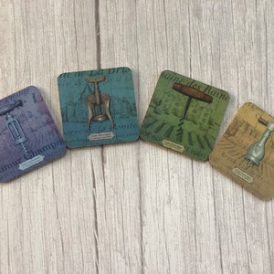 May include: Four square coasters with a vintage design featuring a corkscrew and text in French. The coasters are in shades of blue, green, and brown.