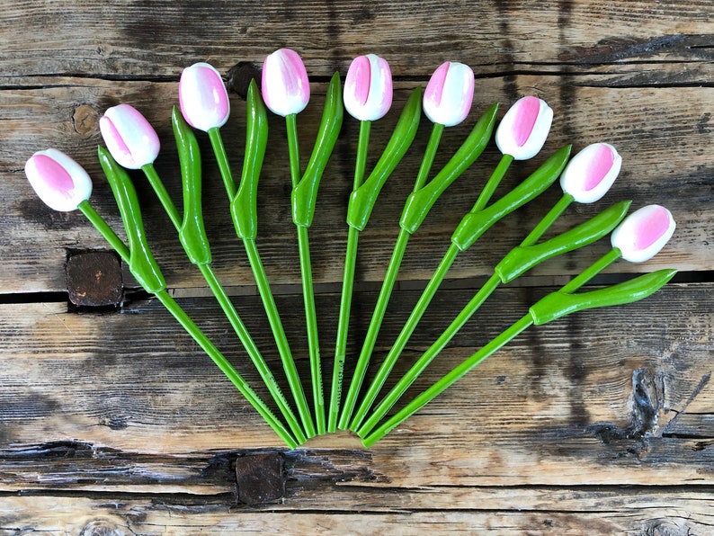 May include: A set of 12 wooden tulips with pink and white painted petals and green stems. The tulips are arranged in a fan shape on a wooden surface.