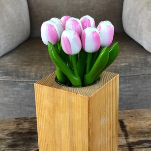 May include: A wooden vase with a square base and a round opening holds a bouquet of pink and white tulips with green leaves.