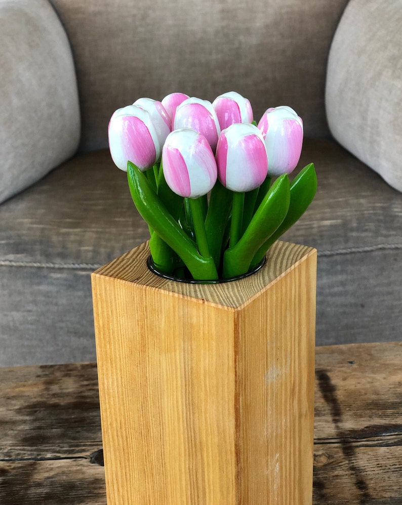 May include: A wooden vase with a square base and a round opening holds a bouquet of pink and white tulips with green leaves.
