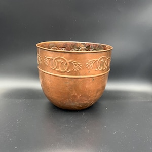 Op de afbeelding: Een koperkleurige kom met een afgeronde basis en een decoratieve band nabij de bovenrand. De band heeft een reliëfontwerp van verweven cirkels en druiventrossen. De kom heeft een gladde, gepolijste afwerking.