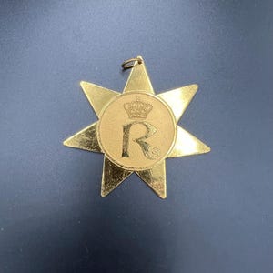 以下が含まれることがあります： 金色の星型のペンダントで、中央は円形です。中央には文字「R」の上に王冠があります。ペンダントの上部には、吊り下げるための小さなループが付いています。ペンダントは濃い青色の表面にあります。