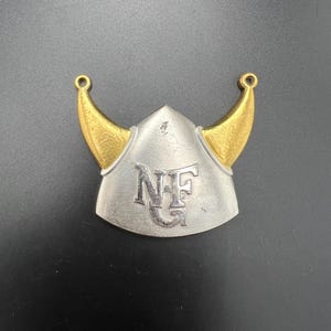 以下が含まれることがあります： 金色の角と中央に「NFG」の文字が入った、銀色のバイキングヘルメット型のペンダント。ペンダントの上部には、チェーンやその他のジュエリーに取り付けるための小さなループが2つあります。