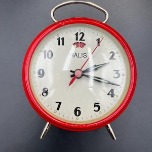 Puede incluir: Un reloj despertador rojo vintage con esfera blanca y números negros. El reloj tiene un asa y patas plateadas, con la palabra "HALIS" impresa en la esfera. Las manecillas del reloj son negras y rojas.