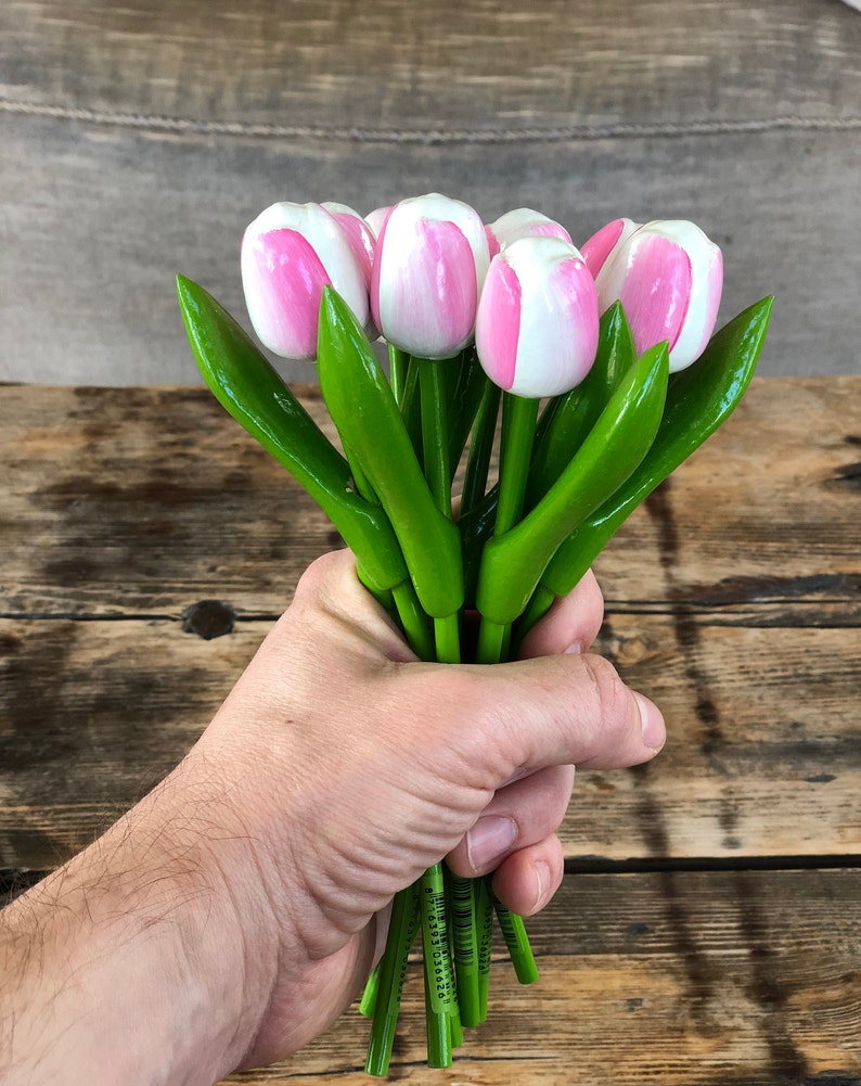May include: A bouquet of wooden tulips with pink and white painted petals and green stems. The tulips are held in a hand.