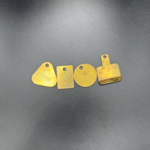 以下が含まれることがあります： さまざまな形状の真鍮製メタルタグ4つ。三角形、長方形、円形、小さなまな板の形。各タグには、吊り下げ用の小さな穴があります。タグは温かみのあるゴールド色で、暗い背景に配置されています。
