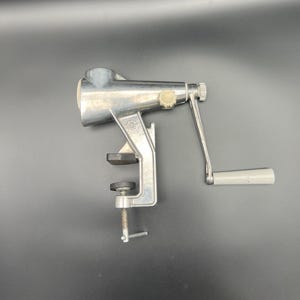 May include: A silver manual citrus juicer with a clamp base and a crank handle. The juicer has a white plastic knob on the crank handle.