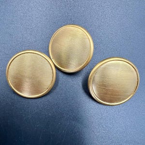 May include: Three gold-coloured, round buttons with a ridged, textured surface. The buttons are arranged on a dark blue surface. These buttons are likely for clothing or fabric items.