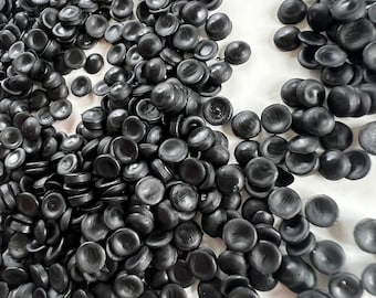 Könnte beinhalten: Eine große Menge schwarzer Kunststoffgranulate, die kleinen Knöpfen ähneln, sind auf einer weißen Oberfläche verstreut.