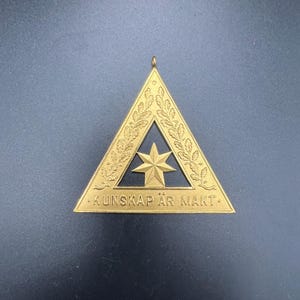 May include: A gold-colored triangular pendant with a star in the center and the text "KUNSKAP ÄR MÅKT" at the bottom. The pendant has an oak leaf design around the edges and a small loop at the top for hanging.