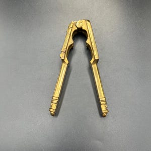 Peut inclure: Un casse-noix en laiton vintage avec un design orné. Le casse-noix est ouvert, révélant son mécanisme à levier. Le laiton a une teinte dorée chaude, et l'outil est posé sur un fond sombre.