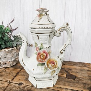 Peut inclure: Une théière en porcelaine blanche avec des accents dorés et floraux. La théière a un couvercle avec un motif floral et une poignée avec un motif enroulé. La théière est décorée de roses tridimensionnelles dans des tons de rose, jaune et orange.