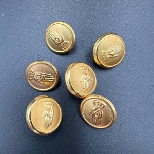 Może przedstawiać: Sześć złotych guzików z wypukłym wzorem dłoni. Guziki są okrągłe z żebrowanym brzegiem. Wzór dłoni jest szczegółowy, z widocznymi palcami i liniami dłoni. Idealne do szycia.