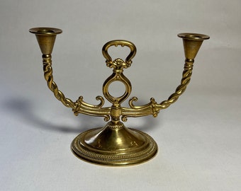 May include: A gold-toned metal candle holder with two arms and a decorative base. The arms are twisted and have a simple, round candle cup at the top. The base is round and has a decorative design.