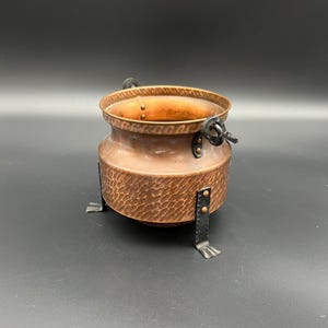 May include: A small copper pot with a hammered finish and black metal legs and handles. The pot has a decorative rim and is ideal for a small plant or decorative display.