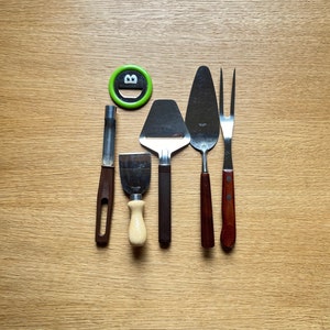 Może przedstawiać: Zestaw przyborów kuchennych, w tym krajalnica do sera, łopatki do ciasta i widelec, z drewnianymi i metalowymi uchwytami. Obecny jest również zielony okrągły magnes z uśmiechniętą twarzą. Przedmioty ułożone są na jasnobrązowej drewnianej powierzchni.