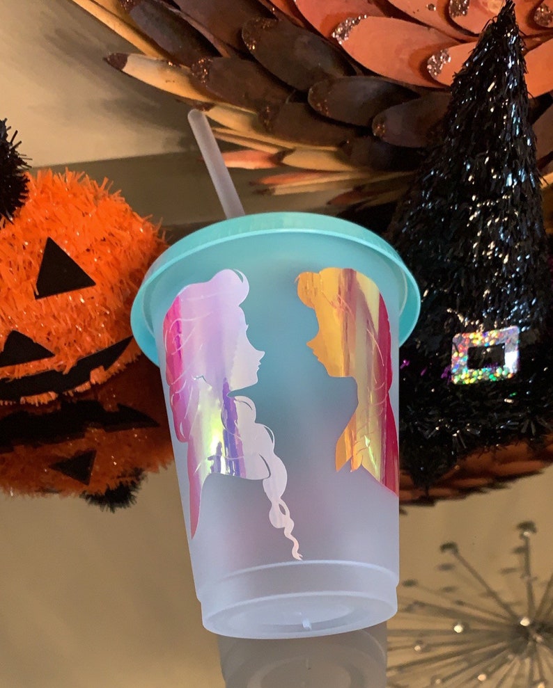 May include: A frosted, light blue reusable cup with a clear plastic straw. The cup has a silhouette of two women with long hair, one with a pink and purple iridescent design and the other with a yellow and orange iridescent design.
