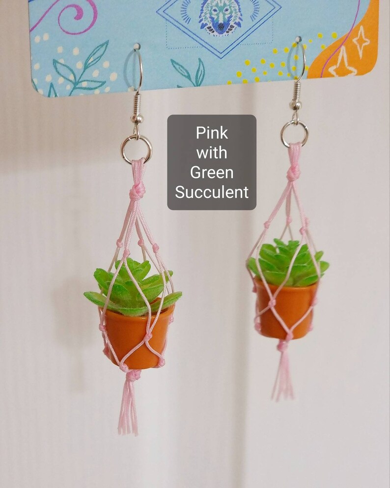 Puede incluir: Pendientes colgantes de macetero rosa con una suculenta verde en una maceta peque&ntilde;a de terracota. Los pendientes est&aacute;n hechos con cuerda rosa y tienen borlas.