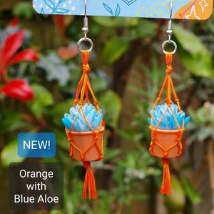 Puede incluir: Un par de pendientes de macram&eacute; naranja con plantas de aloe azul. Los pendientes son nuevos y tienen un gancho plateado.