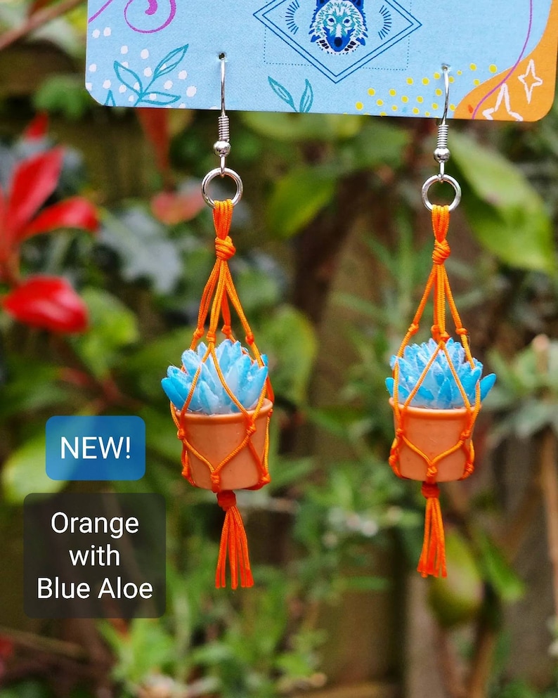 Puede incluir: Un par de pendientes de macram&eacute; naranja con plantas de aloe azul. Los pendientes son nuevos y tienen un gancho plateado.