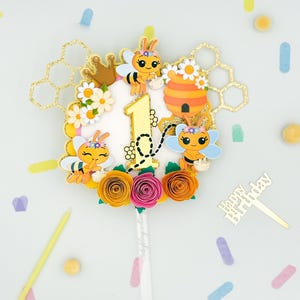 Puede incluir: Un adorno para tarta colorido con tem&aacute;tica de abejas, con un n&uacute;mero 1 dorado, abejas de dibujos animados, una colmena y flores de papel en naranja, rosa y amarillo. El adorno incluye un palillo dorado de "Happy Birthday" y detalles de panal.
