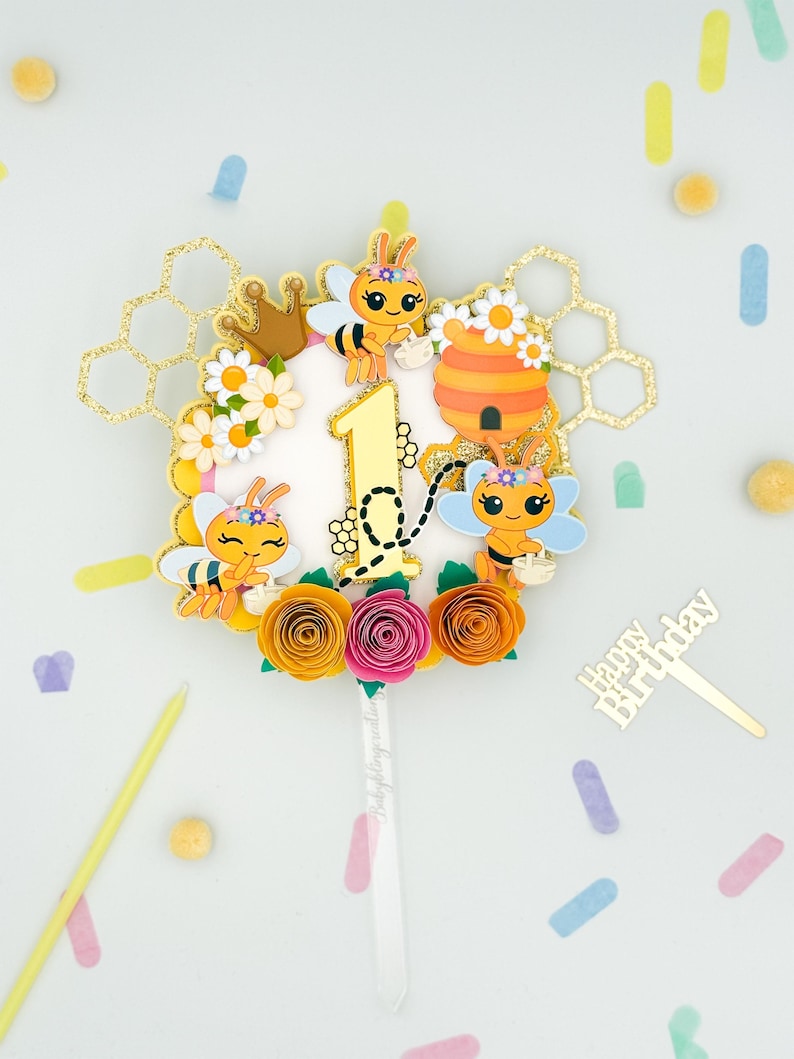 Puede incluir: Un adorno para tarta colorido con tem&aacute;tica de abejas, con un n&uacute;mero 1 dorado, abejas de dibujos animados, una colmena y flores de papel en naranja, rosa y amarillo. El adorno incluye un palillo dorado de "Happy Birthday" y detalles de panal.