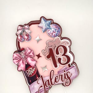 May include: A pink and burgundy cake topper with a Dr. Pepper bottle, cherries, disco ball, star, and the number 13. The name "Jaleris" is written on a banner. The topper is designed for a birthday celebration.