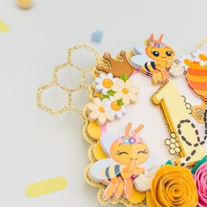 Puede incluir: Adorno para tarta con tem&aacute;tica de abejas, con un dise&ntilde;o de panal dorado con purpurina. Presenta abejas de dibujos animados, flores y el n&uacute;mero "1". El dise&ntilde;o incluye elementos amarillos, naranjas, rosas y blancos, ideal para un primer cumplea&ntilde;os.