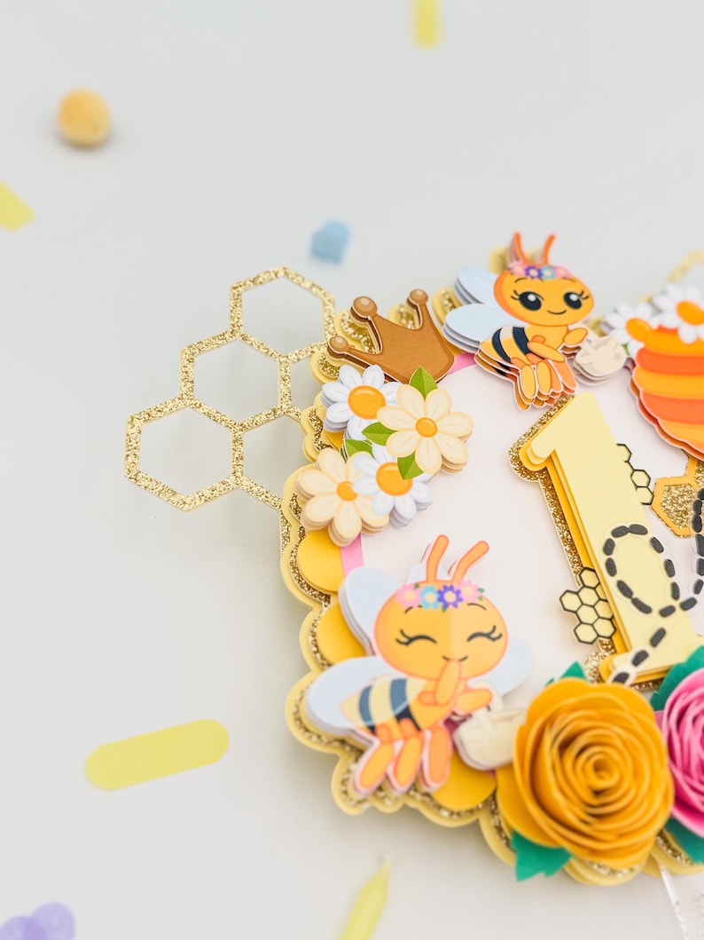 Puede incluir: Adorno para tarta con tem&aacute;tica de abejas, con un dise&ntilde;o de panal dorado con purpurina. Presenta abejas de dibujos animados, flores y el n&uacute;mero "1". El dise&ntilde;o incluye elementos amarillos, naranjas, rosas y blancos, ideal para un primer cumplea&ntilde;os.