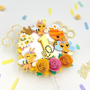 Puede incluir: Adorno para tarta con tem&aacute;tica de abejas. El adorno presenta un n&uacute;mero uno amarillo, abejas de dibujos animados, flores de papel y formas de panal. El adorno est&aacute; decorado con purpurina dorada y est&aacute; dise&ntilde;ado para un primer cumplea&ntilde;os.