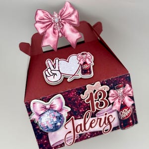 May include: A burgundy gift box with pink accents, featuring a large pink bow on top. Embellishments include a disco ball, a peace sign, a heart, and a Dr. Pepper bottle. The box is decorated with the number "13" and the name "Jaleris".