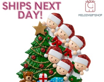 May include: A Christmas tree ornament featuring a family of five peeking around a white picket fence. The ornament is decorated with a green Christmas tree, red bows, and a gold star. The ornament also includes a teddy bear and a wrapped gift.