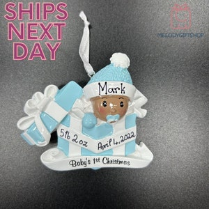 Op de afbeelding: Blauwe en witte kerstversiering met een babyjongetje dat uit een cadeaudoos kijkt. De versiering is gepersonaliseerd met de naam "Mark", de datum "April 4, 2022", en de tekst "5 lb 2 oz" en "Baby's 1st Christmas".