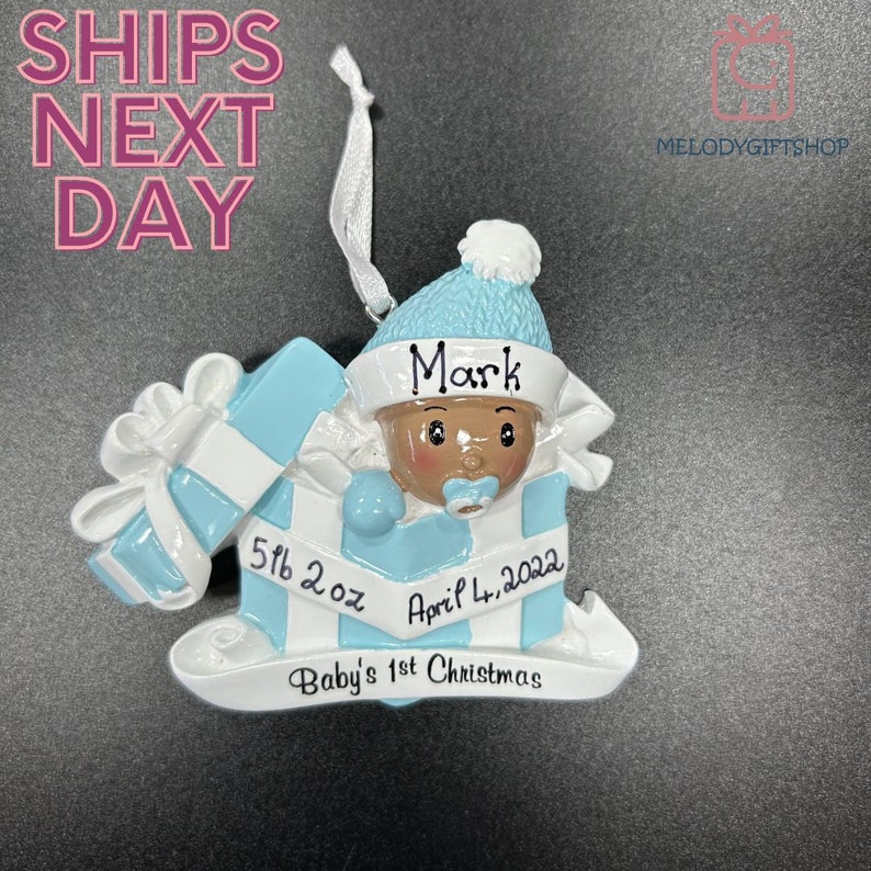 Op de afbeelding: Blauwe en witte kerstversiering met een babyjongetje dat uit een cadeaudoos kijkt. De versiering is gepersonaliseerd met de naam "Mark", de datum "April 4, 2022", en de tekst "5 lb 2 oz" en "Baby's 1st Christmas".