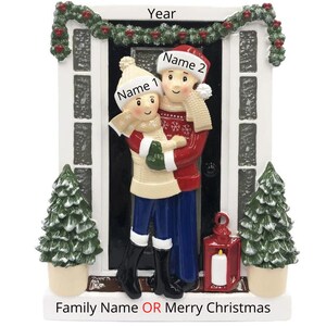 May include: A Christmas ornament featuring a couple standing in front of a door with a wreath. The couple is wearing winter clothes and holding each other. The ornament is decorated with a red lantern and two small pine trees. The text "Year" is at the top of the ornament, and the text "Family Name OR Merry Christmas" is at the bottom.