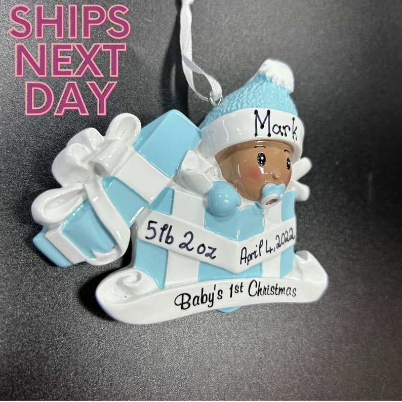 Op de afbeelding: Blauw en wit baby's eerste kerst ornament met een babyjongen die een blauwe muts en een speen draagt. De ornament heeft de tekst "Mark", "5 lb 2 oz", "April 4, 2022", en "Baby's 1st Christmas".