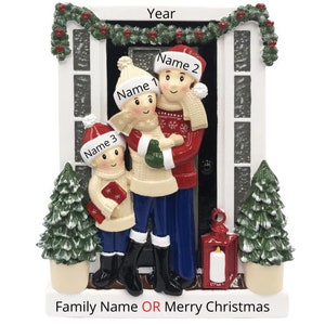 May include: A Christmas ornament featuring a family of three standing in front of a house. The family is wearing winter clothes and the house is decorated with a wreath and garland. The ornament is white with red and green accents. The text "Family Name OR Merry Christmas" is printed on the bottom of the ornament.