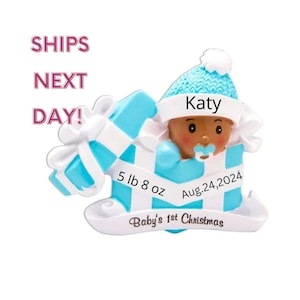 May include: Blue and white personalized baby's first Christmas ornament. The ornament features a baby in a blue hat with a pacifier, inside a blue and white gift box. The ornament includes the baby's name "Katy", birthdate "Aug. 24, 2024", and weight "5 lb 8 oz".