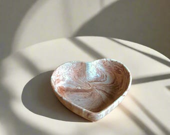 May include: A heart-shaped ceramic dish with a brown and white marbled glaze. The dish is empty and sitting on a light brown surface.
