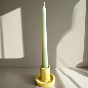 May include: A white candle sits in a yellow marble candle holder. The candle holder has a round base and a cylindrical top.
