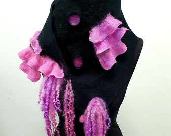 May include: A black felt scarf with pink and purple accents. The scarf features layered pink felt flowers and dangling strands of curly purple and pink wool. The scarf is displayed on a black mannequin.