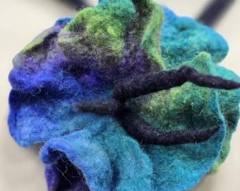 May include: A handmade felt flower in shades of blue, green, and purple. The flower has a textured appearance with dark accents. The felted wool flower is a unique accessory.
