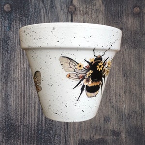 May include: White ceramic flower pot with a black and yellow bee and yellow flowers painted on the side. The pot has a speckled white glaze.